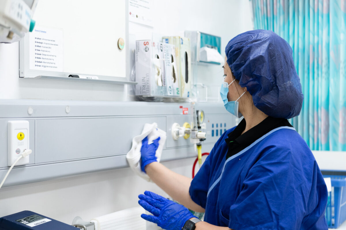 Healthcare cleaner disinfecting high-touch surfaces around a patient care area, ensuring infection control compliance.